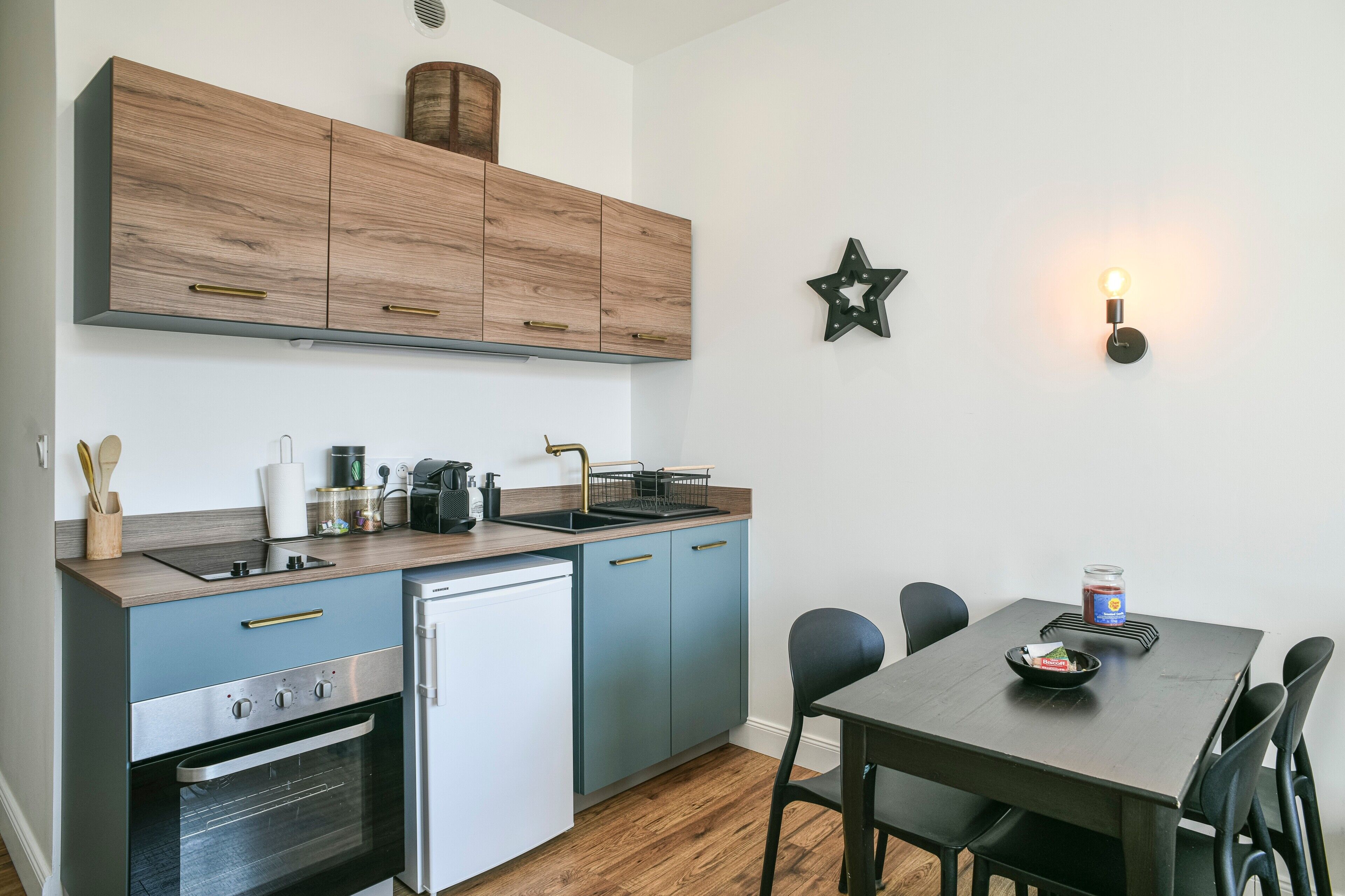 Una cucina compatta con lavandino, fornelli e frigorifero, un tavolo da pranzo con sedie e un lampadario a parete.