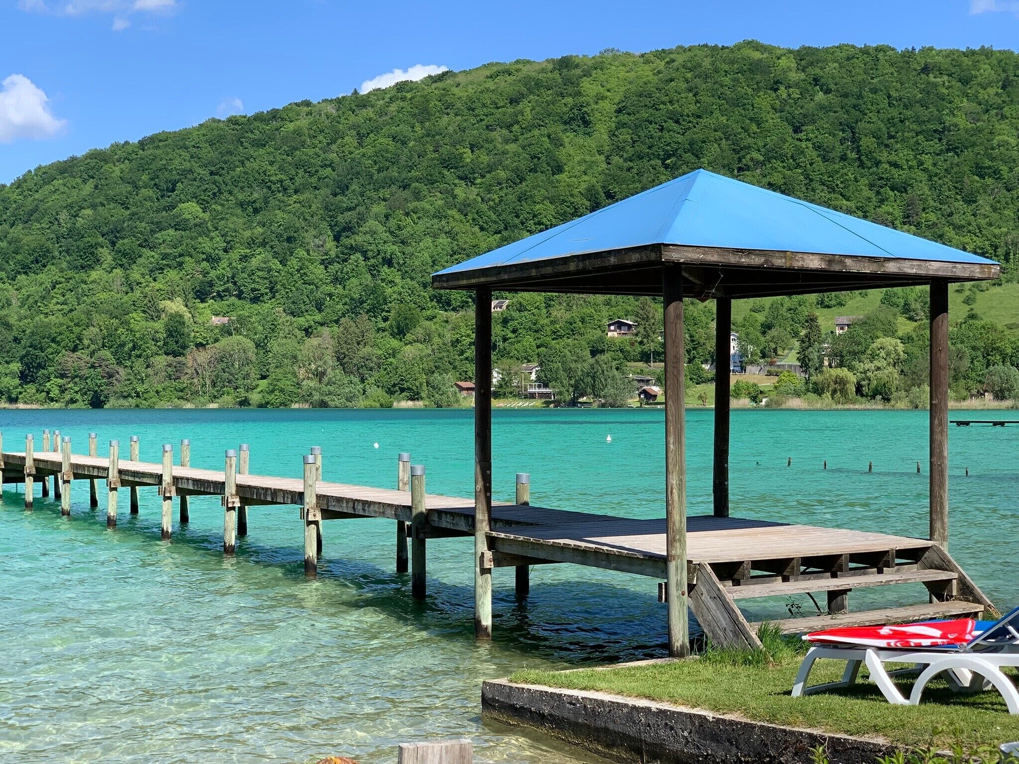 Une jetée en bois s’avance sur un lac aux eaux claires, et à son extrémité se trouve un belvédère surmonté d’un toit bleu.