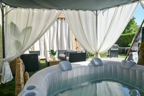 Ein Whirlpool unter einem Baldachin mit weißen Vorhängen, umgeben von Gartenmöbeln und einem Grill.