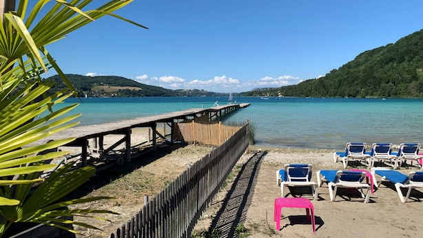 Una playa con un muelle de madera, sillas de descanso y una vista despejada del agua y las colinas al fondo.