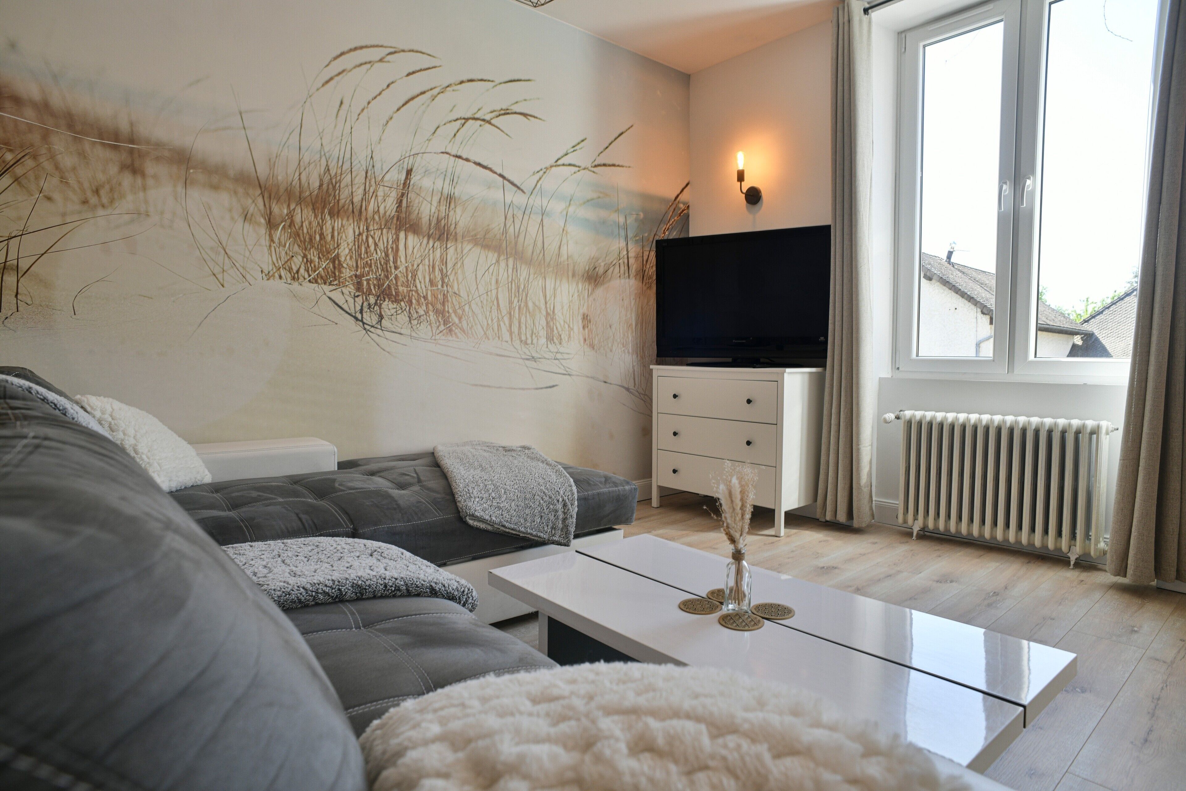 Een moderne woonkamer met een grijze bank, een witte salontafel, een flatscreen televisie en een groot raam met gordijnen.