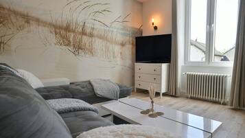Een moderne woonkamer met een grijze bank, een witte salontafel, een flatscreen televisie en een groot raam met gordijnen.