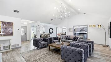 A spacious living room with a large chandelier, a fireplace, and a modern kitchen in the background.
