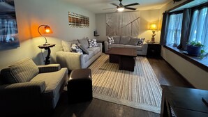 Een woonkamer met een bank, fauteuils, een salontafel, een vloerkleed, een plafondventilator en een raam met gordijnen.