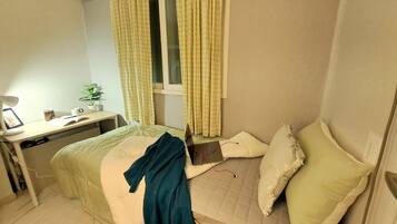 A bedroom with a bed, a desk, a chair, a window with curtains, and a bedside table with a lamp and a plant.