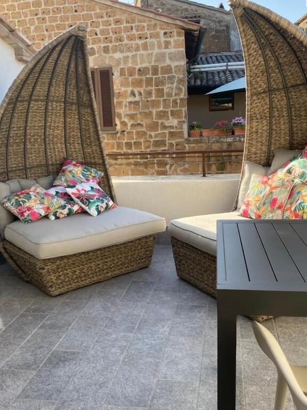 Un espace extérieur accueillant, aménagé avec des meubles en osier et des coussins colorés, adossé à un bâtiment en pierre surmonté d’un toit en tuiles.