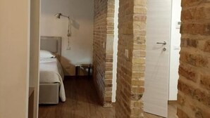 Ein schmaler Flur mit freiliegenden Backsteinwänden und Holzdielen, der zu einem Schlafzimmer mit Bett und Nachttisch führt.