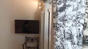 Ein Zimmer mit einem an der Wand befestigten Flachbildfernseher, einem gemusterten Sessel und einem Vorhang mit einer Landschaftsdarstellung.