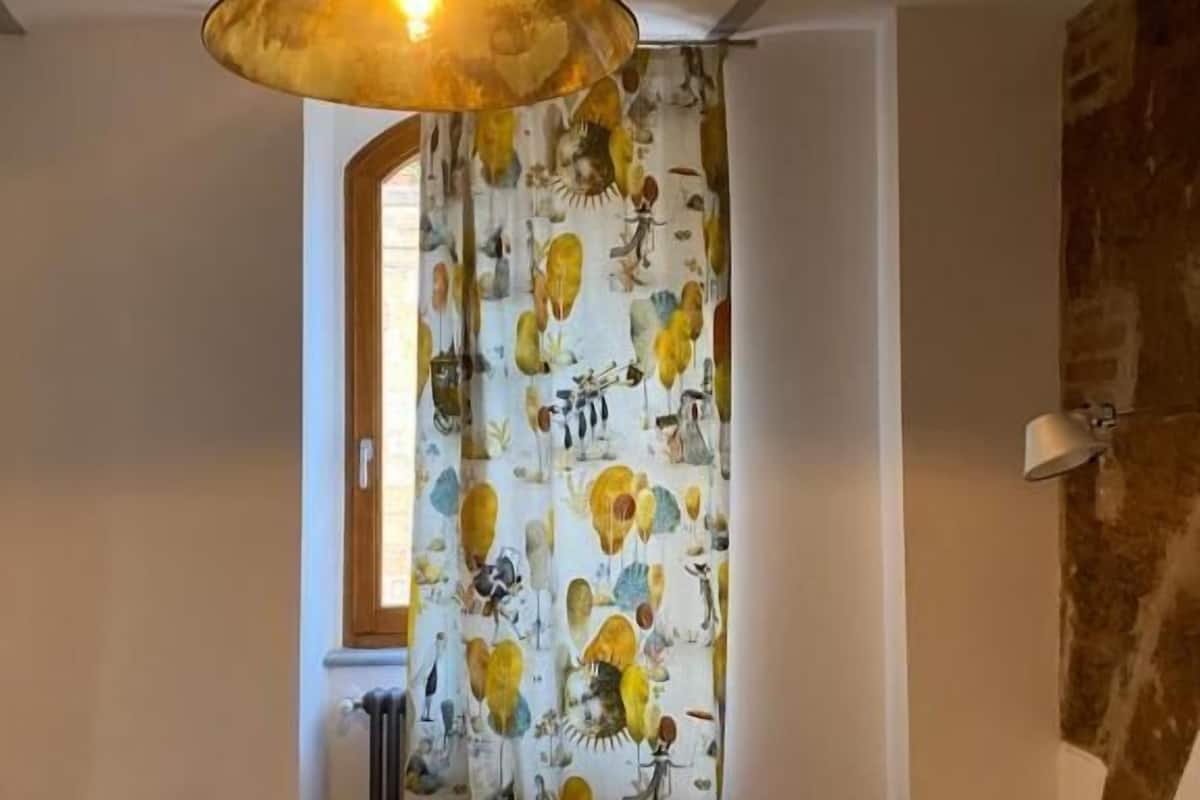 Um quarto com cama, luminária pendente, cortina com padrão de pássaros e radiador.