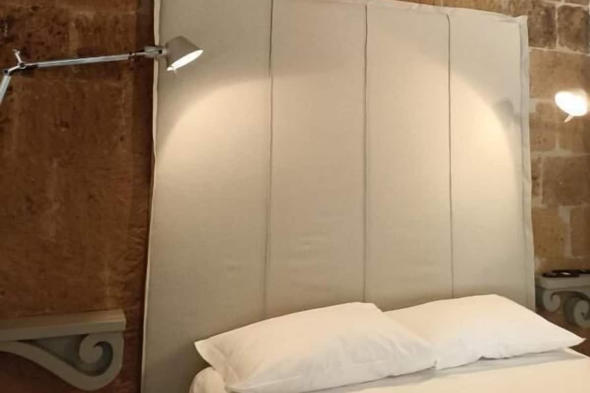 Uma cama com roupa de cama branca, cabeceira e mesa de cabeceira com abajur.