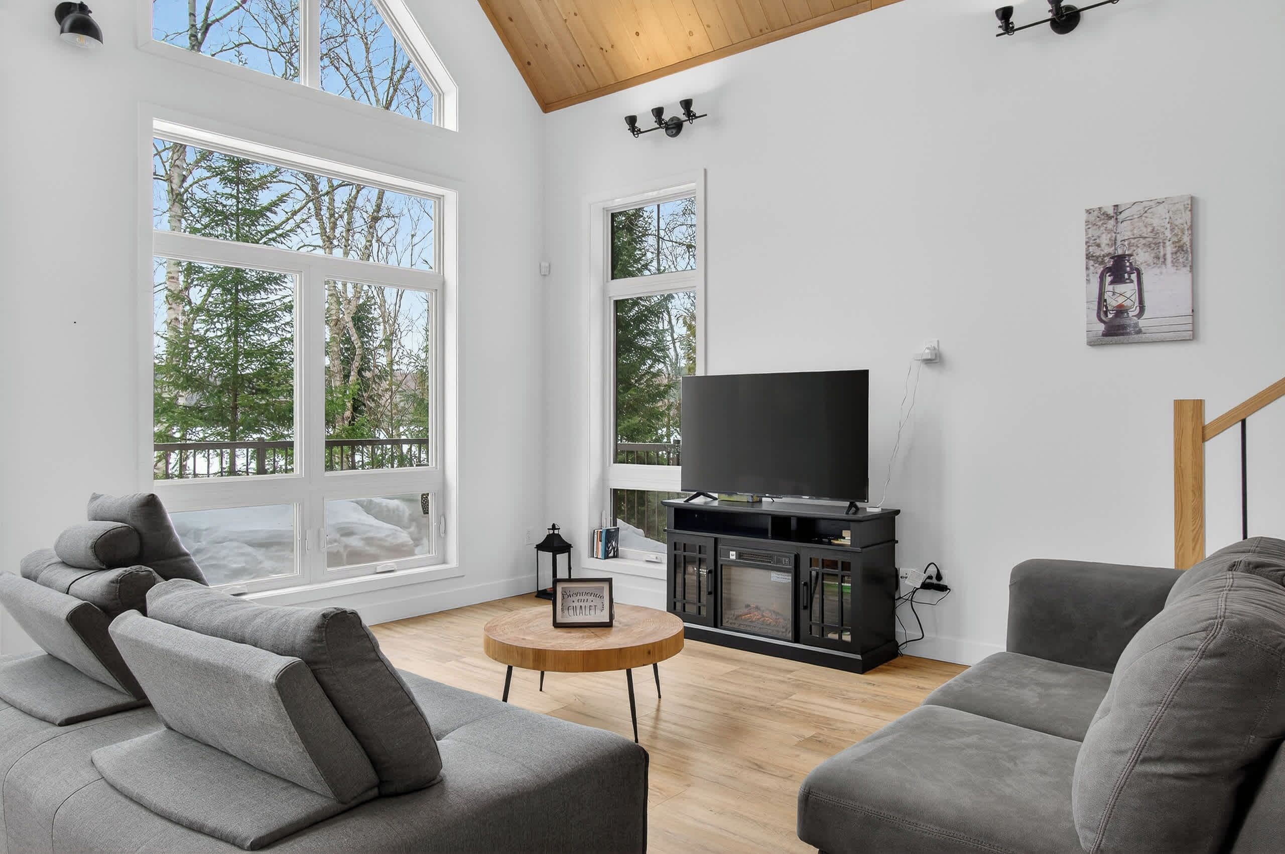 A modern living room with a large TV, grey sofas, a wooden coffee table, and large windows with a view of trees.