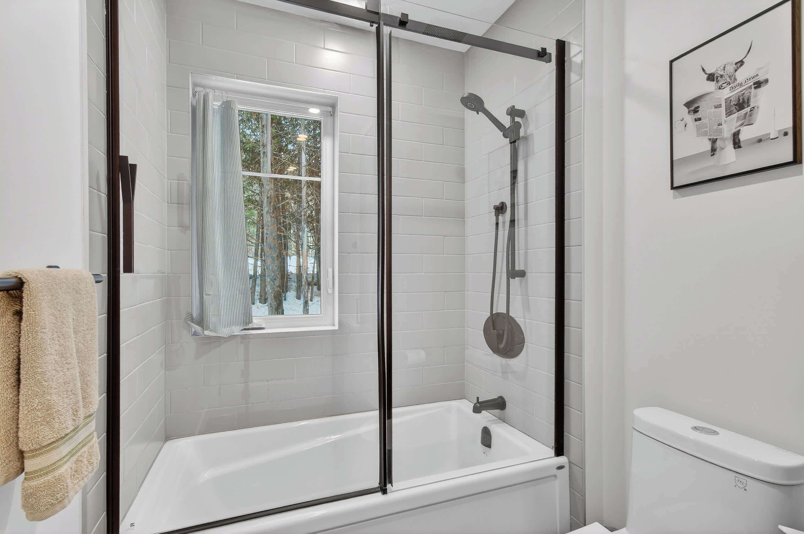 A modern bathroom with a glass-enclosed shower, a white bathtub, and a window with a view of trees.