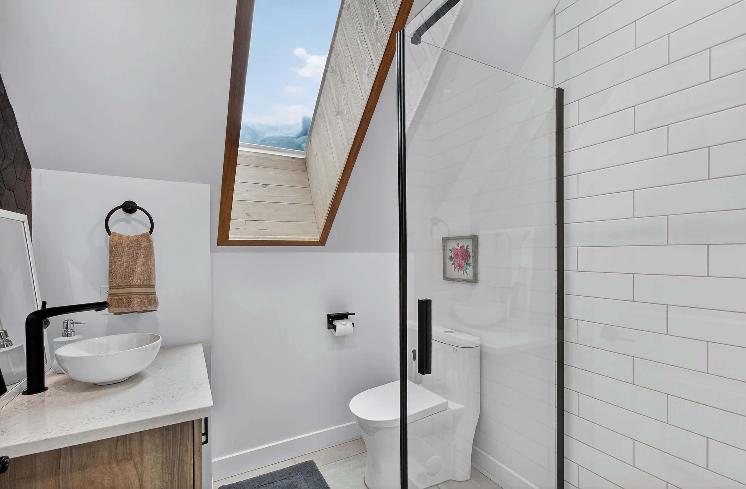 A modern bathroom with a skylight, a white sink, a toilet, and a glass shower enclosure.