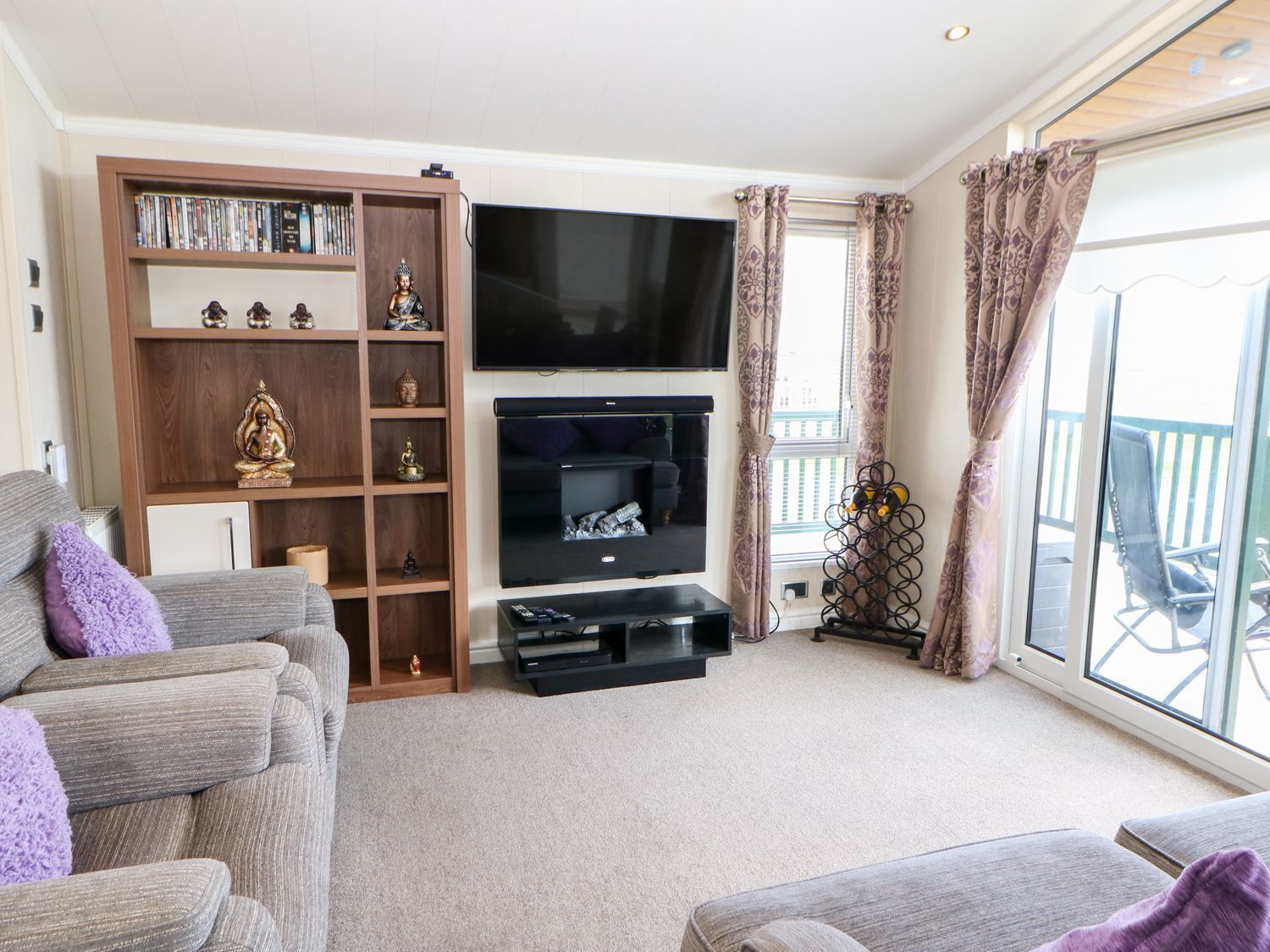 A living room with a flat-screen TV, a bookshelf with decorative items, a grey sofa, and a balcony with a chair.
