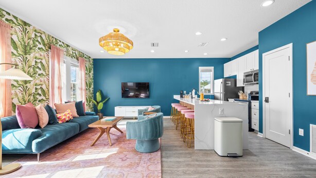 A modern living room with a blue accent wall, a sofa, a coffee table, and a kitchen area with a microwave and bar stools. - A Boho City Escape  Sleeps 8 Tampa Heights (Tampa)