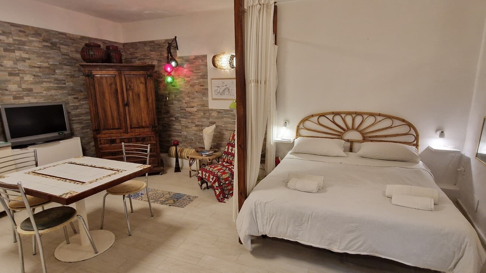 A bedroom with a bed, a wooden headboard, a TV, a desk with chairs, and a patterned rug.