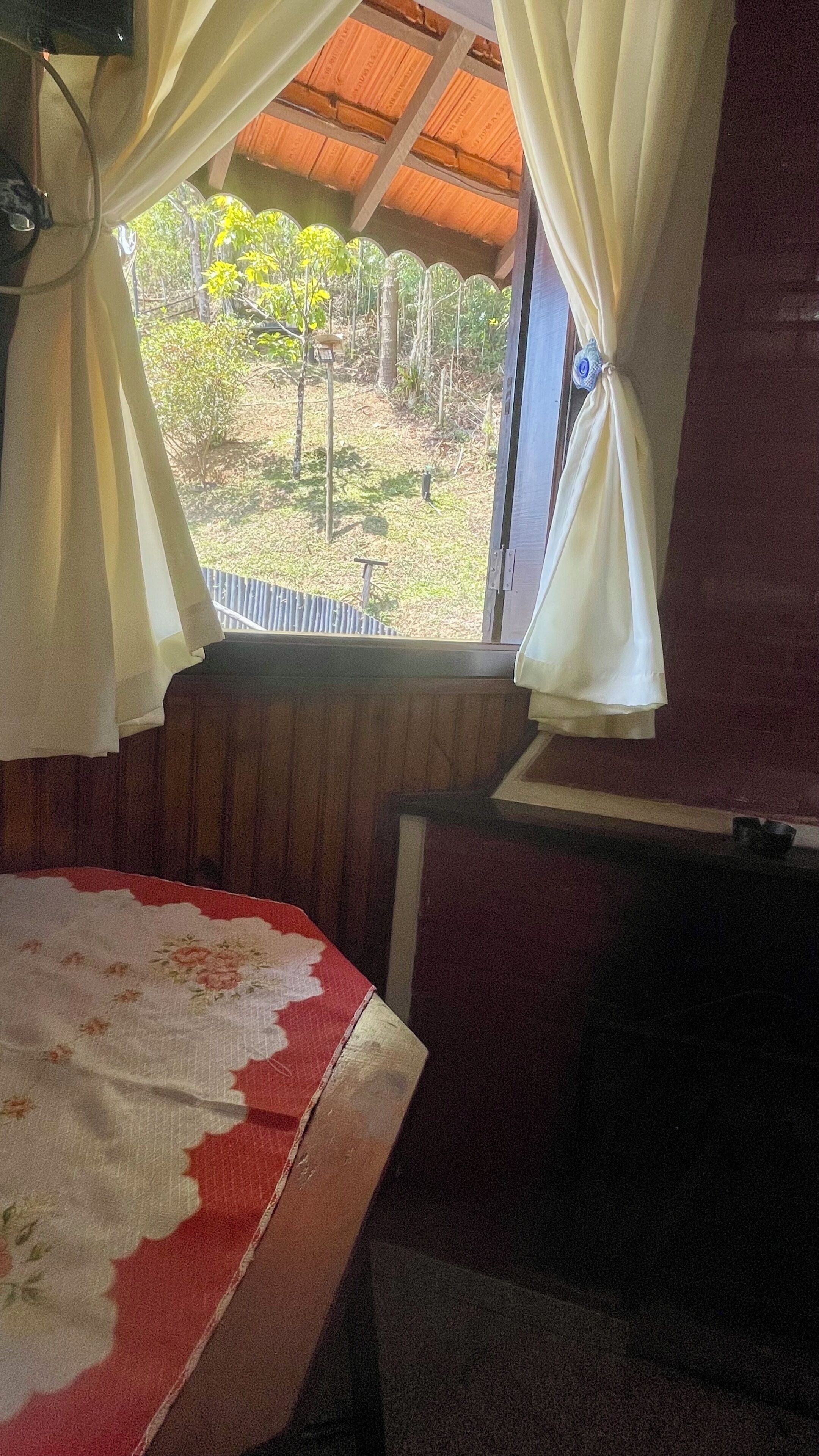 A room with a bed, a window with curtains, a view of greenery outside, and a wooden ceiling.