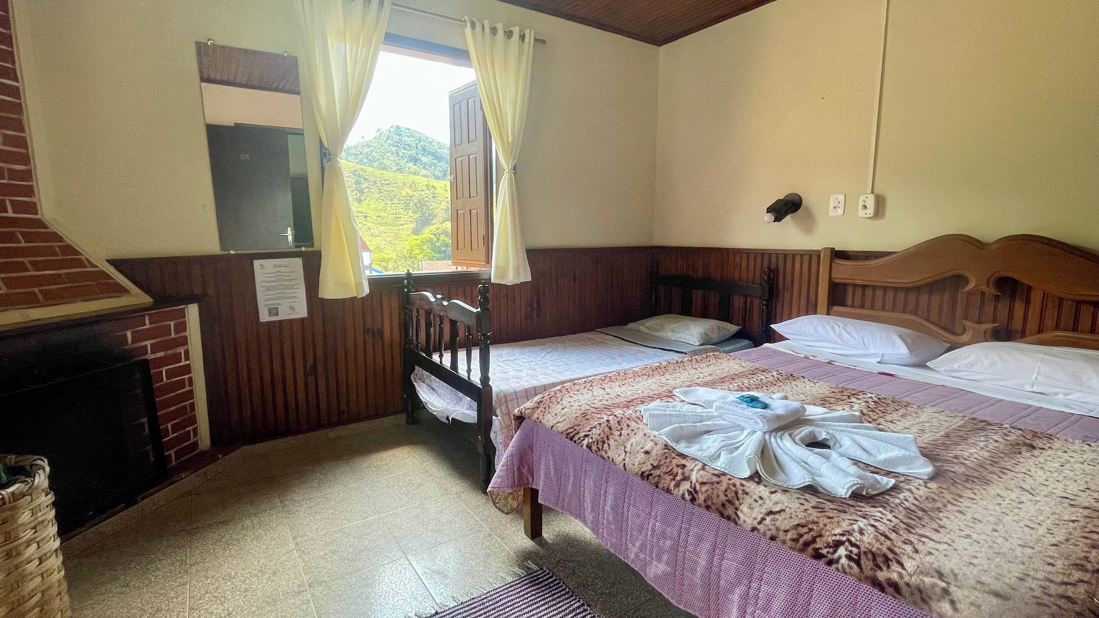 Un dormitorio con cama, cabecera de madera, ventana con cortinas, chimenea y piso de baldosas.