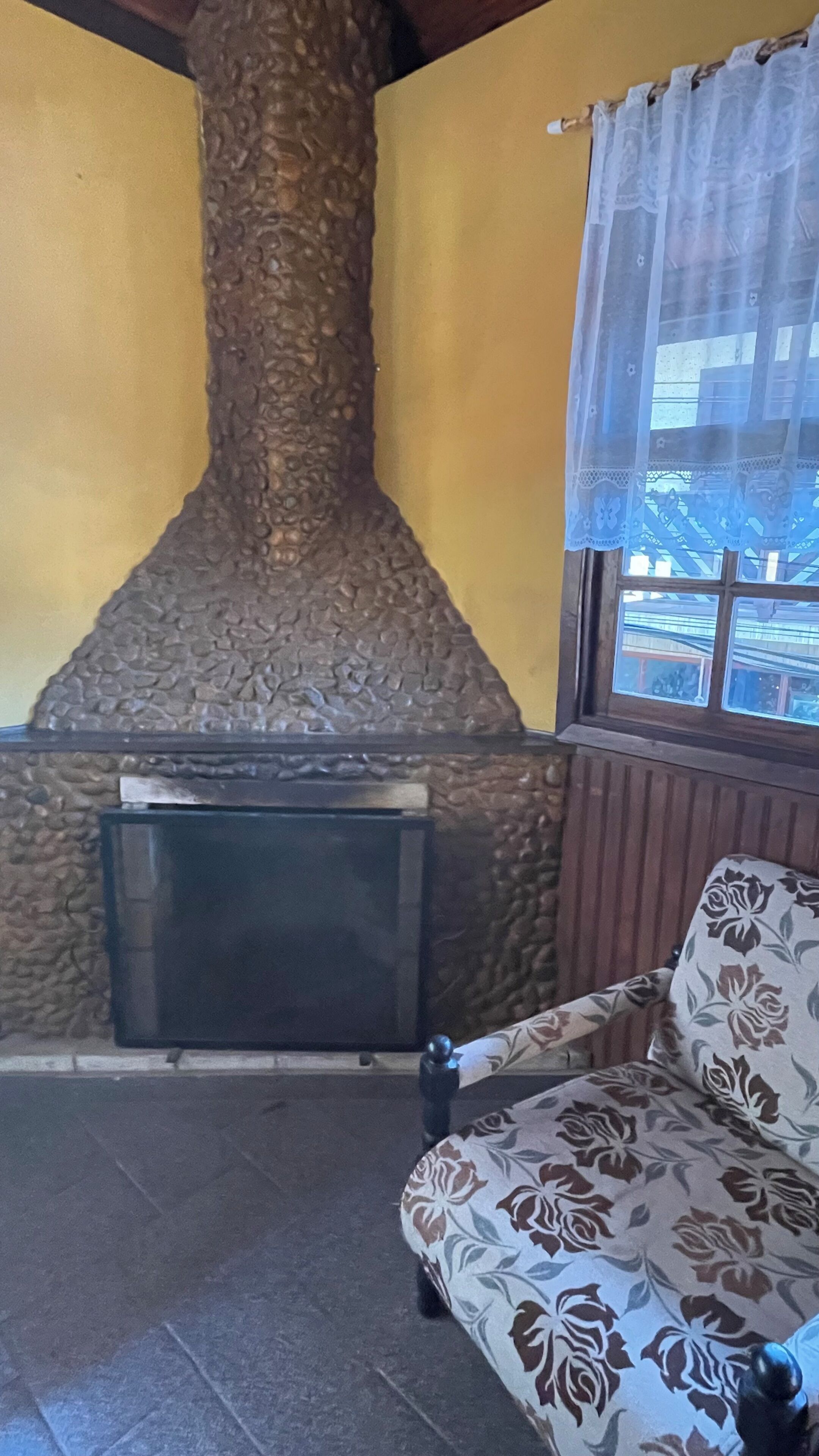A stone fireplace, a sofa with floral upholstery, and a window with lace curtains.