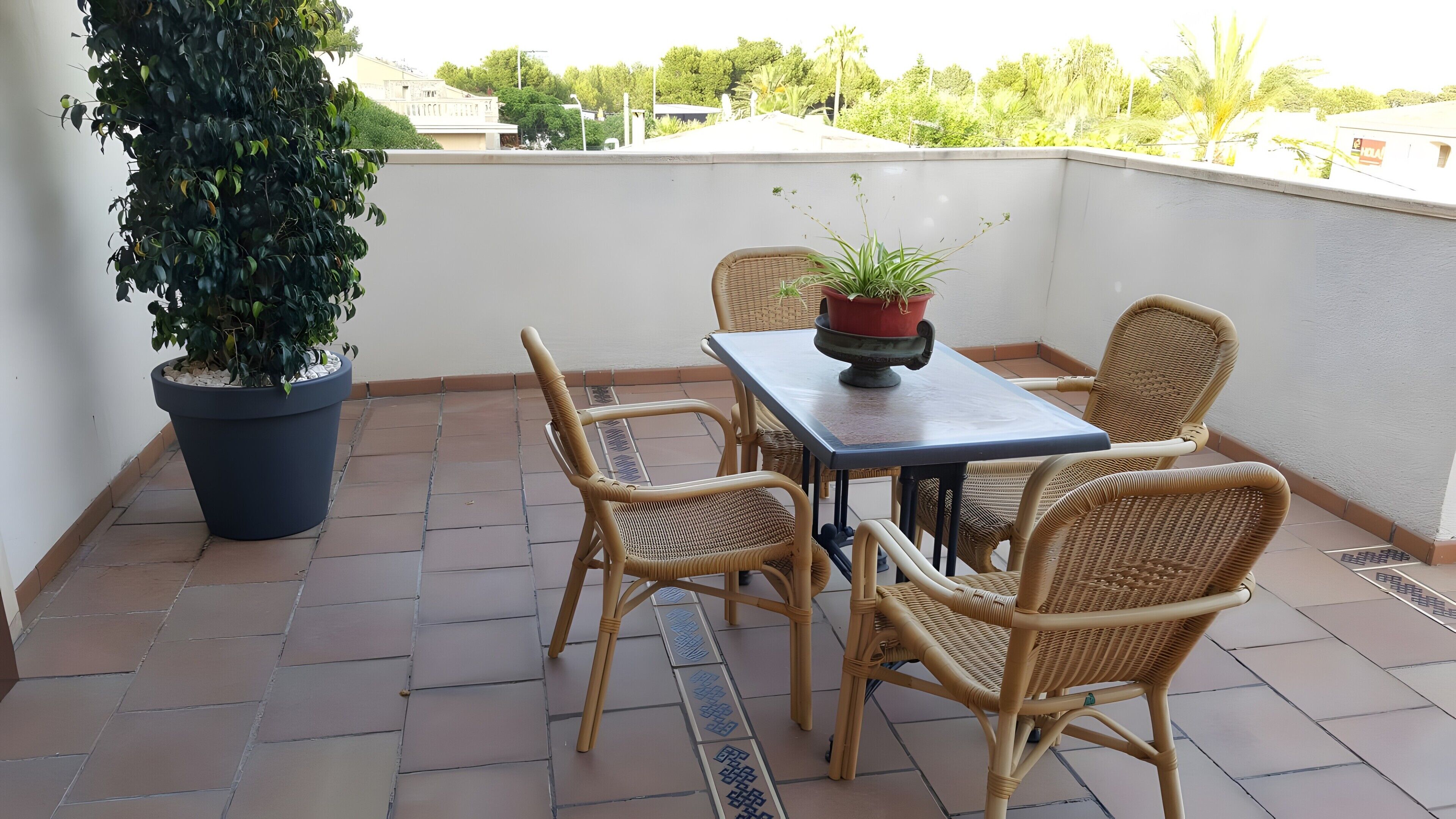 Ein Balkon mit Tisch, vier Korbstühlen, einem Blumentopf und Blick auf Gebäude und Bäume.