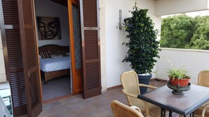 An open balcony with a bed, wicker chairs, a table, and potted plants.