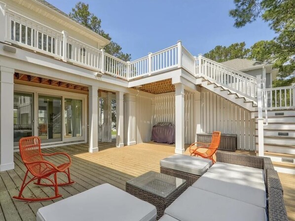 Eine überdachte Terrasse im Freien mit Holzdeck, weißen Geländern und Sitzgelegenheiten, darunter Korbs Sofas und Stühle.