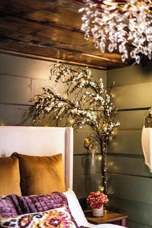 Ein Schlafzimmer mit einem Bett, einem Nachttisch, einer Vase mit Blumen und einem wandmontierten Spiegel.