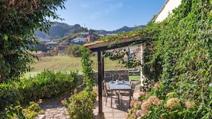A patio with a table and chairs, surrounded by greenery and a view of a hillside town.