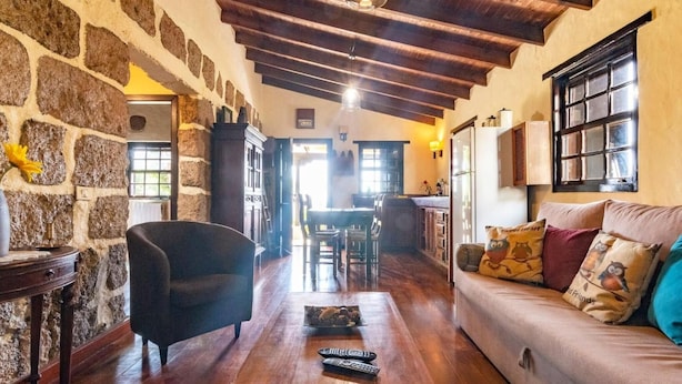 A cozy living room with stone walls, wooden flooring, a sofa with patterned cushions, a coffee table with remotes, and a dining area in the background.