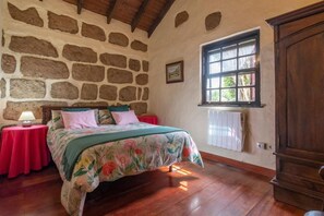 Een slaapkamer met een stenen muur, een bed met bloemenbedrukte lakens, een rode tafel met een lamp, en een raam met uitzicht.
