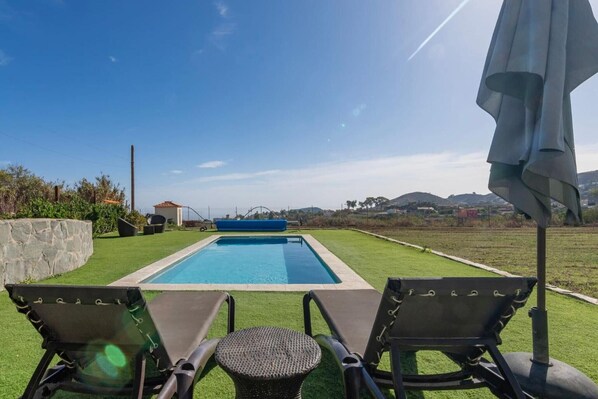 A swimming pool with two lounge chairs and a table, surrounded by a well-maintained lawn and a stone wall.