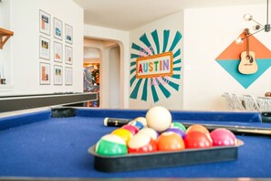 Ein Billardtisch mit bunten Kugeln, eine gerahmte Bildergalerie an der Wand und ein Neon-Schild mit der Aufschrift 'AUSTIN'.