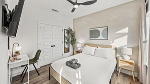 Una camera d'albergo con un letto, una scrivania, una sedia, una TV, una ventola a soffitto, un comodino con una lampada e un'opera d'arte appesa al muro.