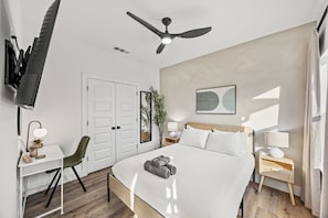 Ein Hotelzimmer mit einem Bett, einem Schreibtisch, einem Sessel, einem Fernseher, einem Deckenventilator, einem Nachttisch mit Lampe und einem Wandbild.