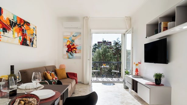 Una sala de estar moderna con comedor, sofá, televisor y balcón con vistas a árboles y edificios.