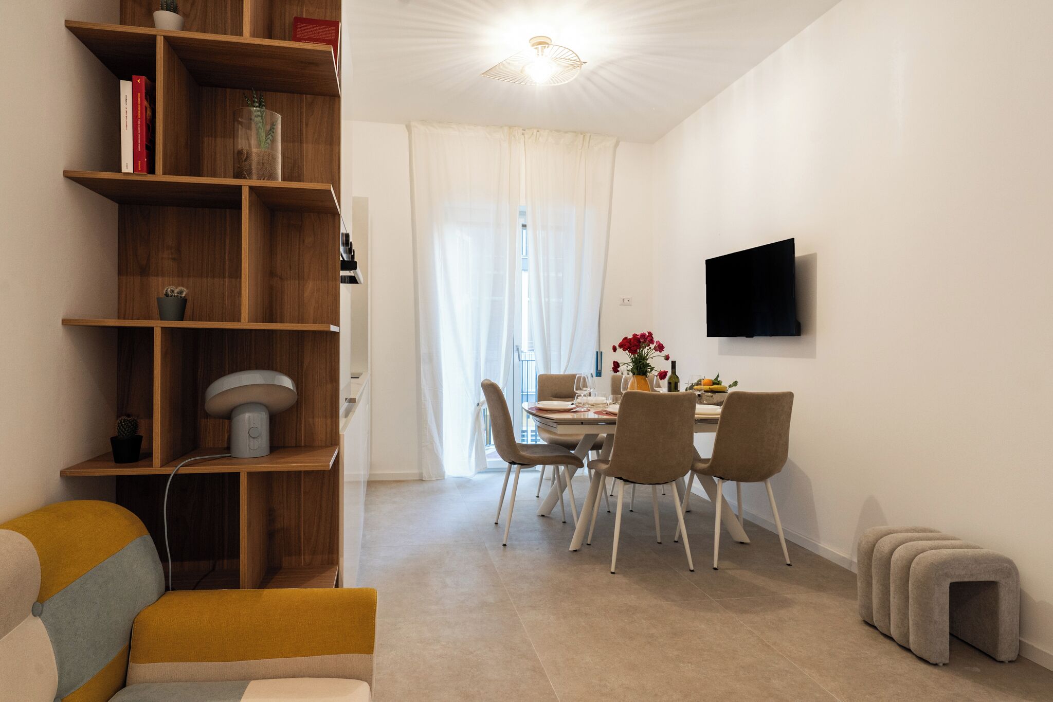 A modern living room with a wooden bookshelf, a dining area with a table and chairs, a flat-screen TV mounted on the wall, and a small stool.