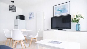Ein modernes, minimalistisches Wohnzimmer mit einem weißen Tisch, zwei weißen Stühlen, einem Flachbildfernseher, einer kleinen Kommode und einer blauen Vase mit weißen Blumen.