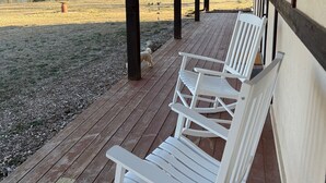 A covered porch with white Adirondack chairs, a wooden deck, and a view of a park with trees and a body of water.