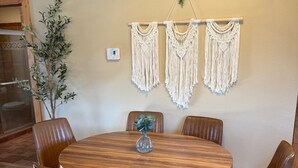 A round wooden table with six brown leather chairs, a vase with a plant, and three macramé wall hangings.