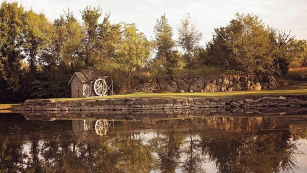 A rustic watermill structure by a calm pond, surrounded by trees and stone walls.