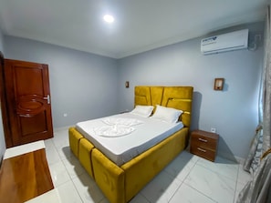 Un dormitorio con una cama amarilla, ropa de cama blanca, una puerta de madera, una mesita de noche pequeña y un equipo de aire acondicionado en la pared.