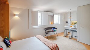 Une chambre à coucher comprenant un lit, une table de chevet, une chaise et un petit bureau avec une autre chaise. Il y a une fenêtre avec des rideaux, un luminaire fixé au mur et une porte.
