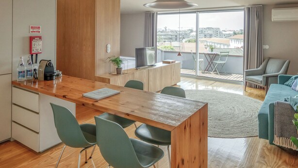 A modern kitchen with a wooden countertop, green chairs, and a dining table. There is a large window with a view of buildings outside. - Penthouse with Private Balcony | Baumhaus Serviced Apartments | Central Porto (Porto)