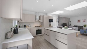 A modern kitchen with white cabinetry, a central island, and a dining area with a grey sofa and a TV. - Modern 3 Bedroom, 2 Bathroom Home in Edgware (Edgware)