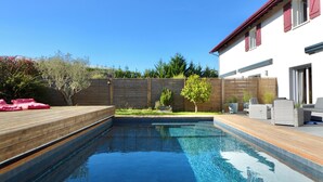 Ein Schwimmbecken mit Holzterrasse, ein weißes Gebäude mit roten Fensterläden und ein klarer, blauer Himmel.