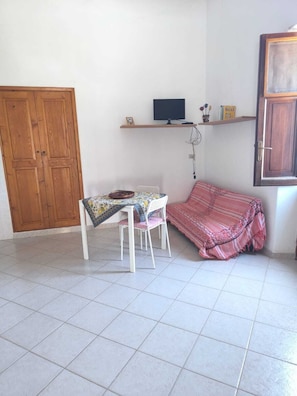 Una habitación con piso de baldosas, una silla blanca, una mesa con mantel estampado, un sofá a rayas rojo y blanco, una puerta de madera y un estante con un televisor y objetos decorativos.
