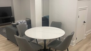 A modern dining area with a round table and four grey chairs, a sofa, and a TV in the background. - Spacious Basement Apartment in Rouge Hill Near Highway 401 (Toronto)