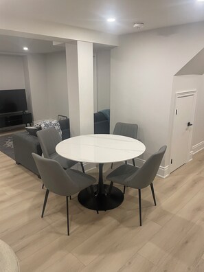 Un espace repas moderne avec une table ronde et quatre chaises grises, un canapé et un téléviseur en arrière-plan.