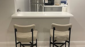 A modern kitchen with a white countertop, stainless steel appliances, and two bar stools. - Spacious Basement Apartment in Rouge Hill Near Highway 401 (Toronto)