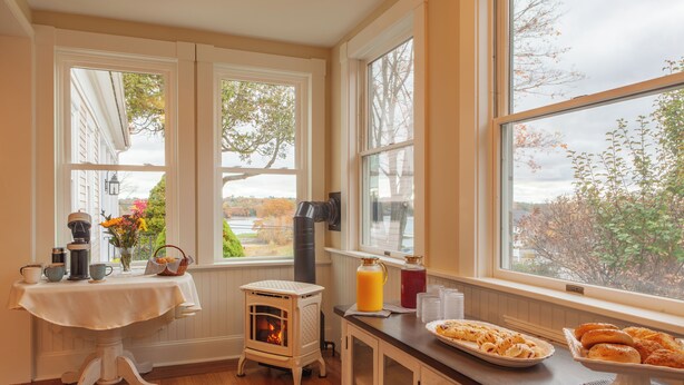 A cozy kitchen with a wood stove, a table with breakfast items, and large windows with a view of trees.